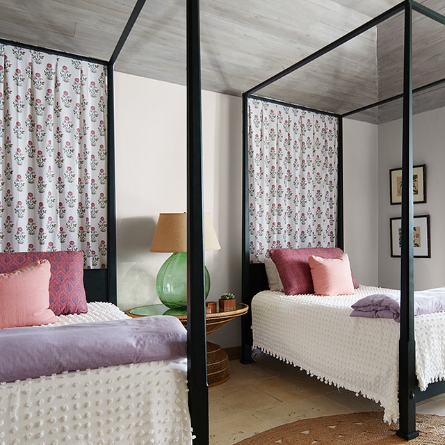 Two black canopy twin beds fit nicely in this off-white painted bedroom with a vaulted shiplap ceiling, colorful pastel and white décor, and a door open to the outside.