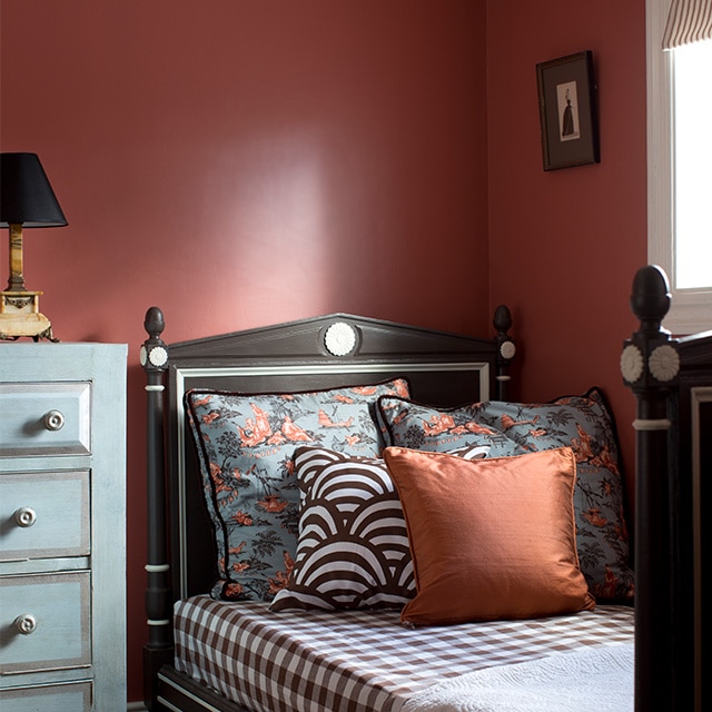 An inviting corner bedroom space with warm red painted walls, a blue dresser and a single bed with eclectic print bedding underneath a white trimmed window.