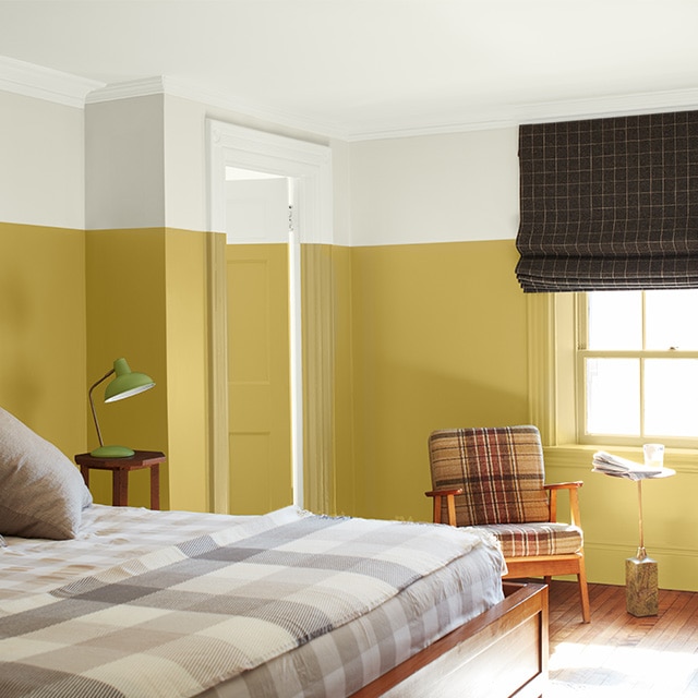 A bright two-tone painted bedroom with white-painted upper walls, upper door and ceiling, yellow-painted lower walls and door, tan checked bedding and two plaid chairs.