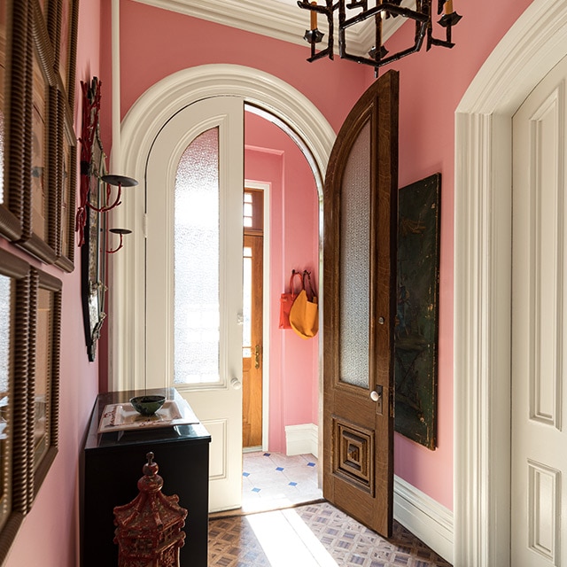 A traditional hallway and vestibule with pink-painted walls, white trim, doors, and ceiling, a bureau and chandelier.