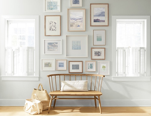 An off-white-painted wall with a collection of framed art behind a wooden bench and shopping bags.