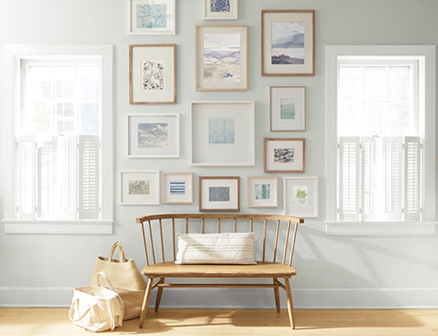 An off-white painted wall with a collection of framed art behind a wooden bench and shopping bags.