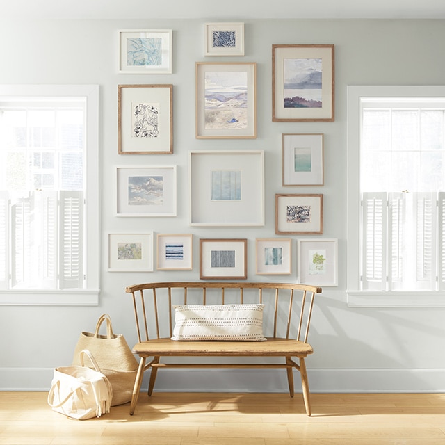 An off-white-painted wall with a collection of framed art behind a wooden bench and shopping bags.