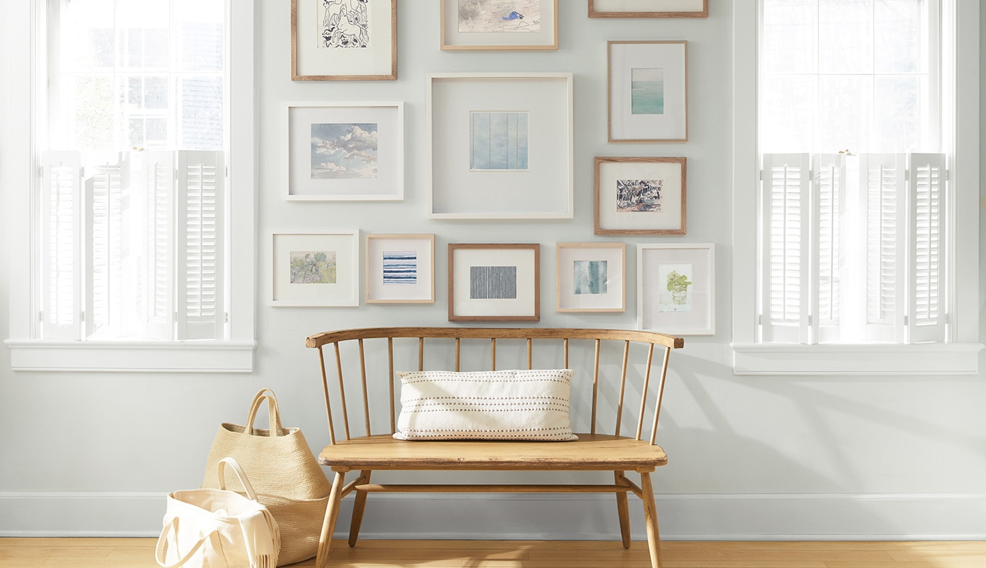 An off-white-painted wall with a collection of framed art behind a wooden bench and shopping bags.