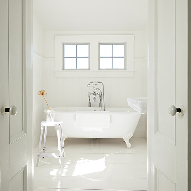 Une salle de bains entièrement peinte en blanc arborant deux fenêtres à moulures blanches au-dessus d’une baignoire sur pieds et des portes et un plafond peints en blanc.