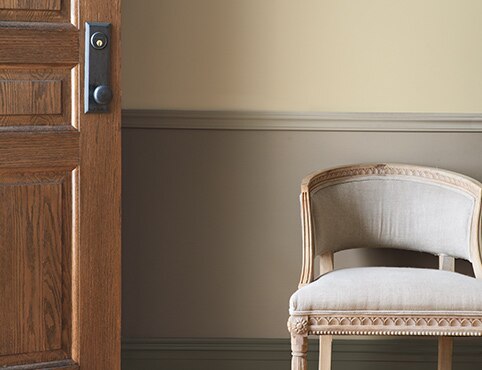 Taupe entryway with neutral-painted trim and wainscoting, wooden door, and upholstered beige chair.