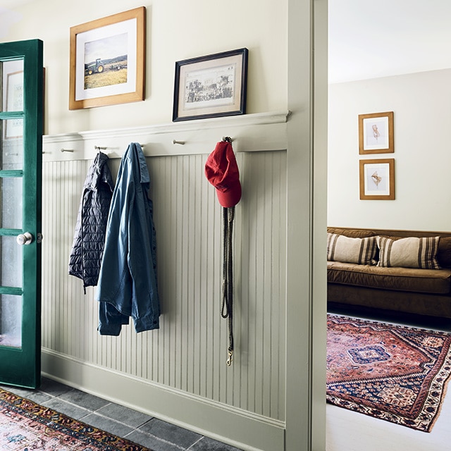 A sunlit entry hallway with a dark green front door, off-white walls, pale green painted wainscotting and trim, and a side room in neutral tones seen through an open doorway.