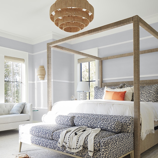 A bright and airy light-gray painted bedroom with white wall trim and ceiling, a modern four-poster bed, a fun cheetah animal print sofa, and wicker chandelier.