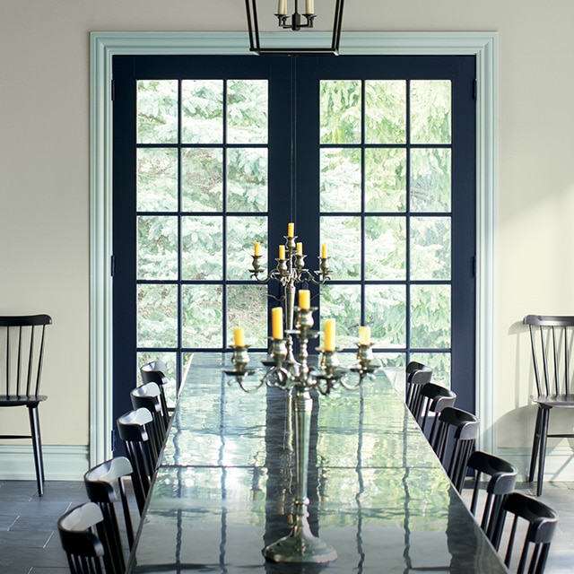Beautiful dark navy blue-painted French doors with blue-gray trim stand out in this sunlit cool gray-painted dining room with a long black table and chairs.