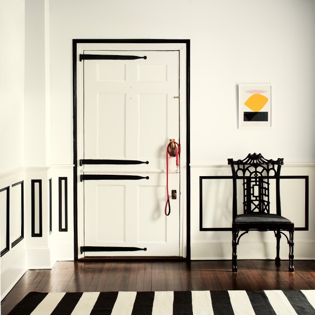This black-and-white styled room features white-painted walls, ceiling and door with black trim and black decorative hinges on the door, a black chair and black-and-white striped rug. 