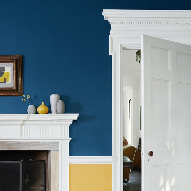 A striking two-tone painted living room wall with navy blue on the top, yellow on the bottom, beautiful white trim and moulding, fireplace mantel and open door.
