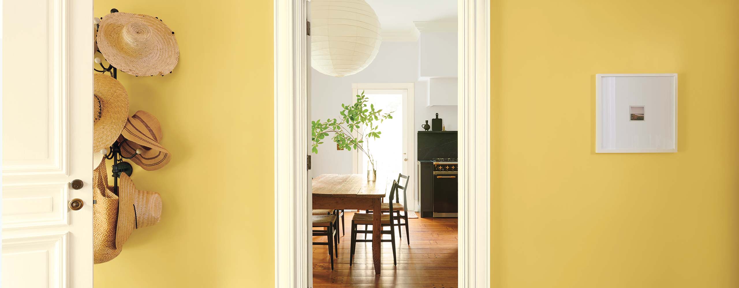 A pretty golden yellow-painted entryway with a mounted hat rack, and a white open door and trim, looking into a light gray-painted kitchen. 