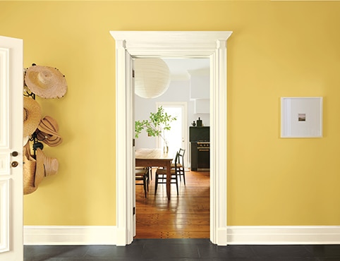 A pretty golden yellow-painted entryway with a mounted hat rack, and a white open door and trim, looking into a light gray-painted kitchen. 