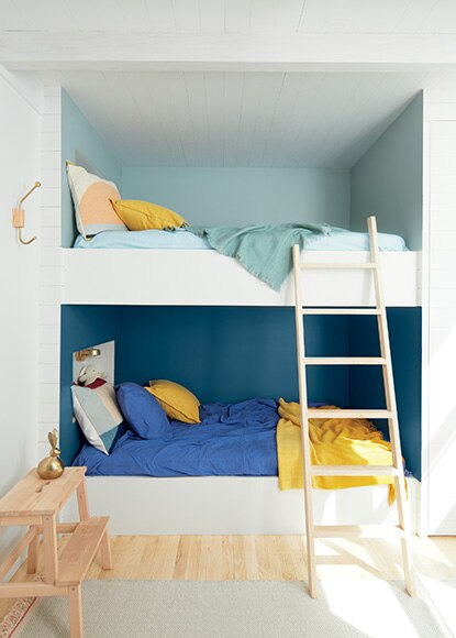 White bedroom with alcove bunk beds in shades of blue with matching bedding, yellow pillows, wooden stepstool, and ladder.