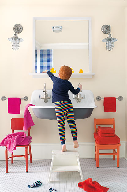 Salle de bains crème avec une chaise orange et une chaise rouge, serviettes assorties, tabouret blanc, luminaires en forme de lanterne et petit garçon s’amusant devant un lavabo gris.