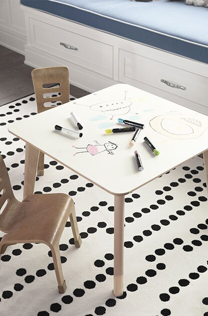 Table dont la surface qui s’essuie à sec avec gribouillis et marqueurs, chaises en bois, tapis à pois et tiroirs blancs surmontés d’un coussin gris.