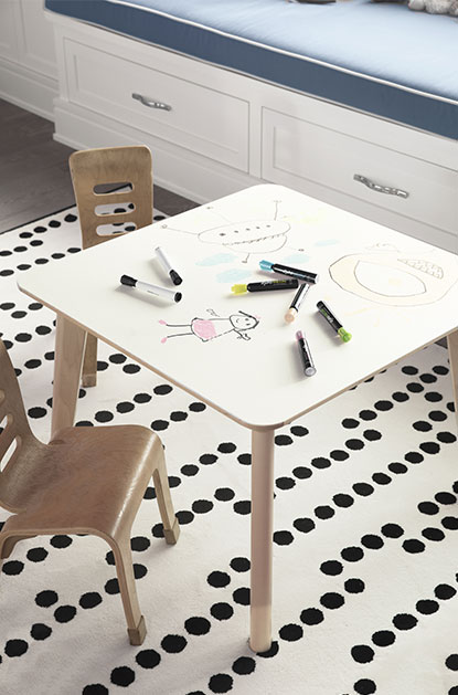 Doodles and markers on a dry-erase tabletop with wooden chairs, a polka-dot-rug, and white drawers topped with gray cushion. Doodles and markers on a dry-erase tabletop with wooden chairs, a polka-dot-rug, and white drawers topped with gray cushion.