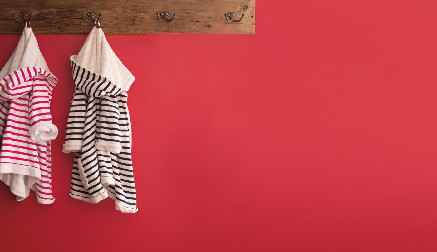 Red wall with a wooden plank coat rack, two striped children's robes, one in pink and one in black.