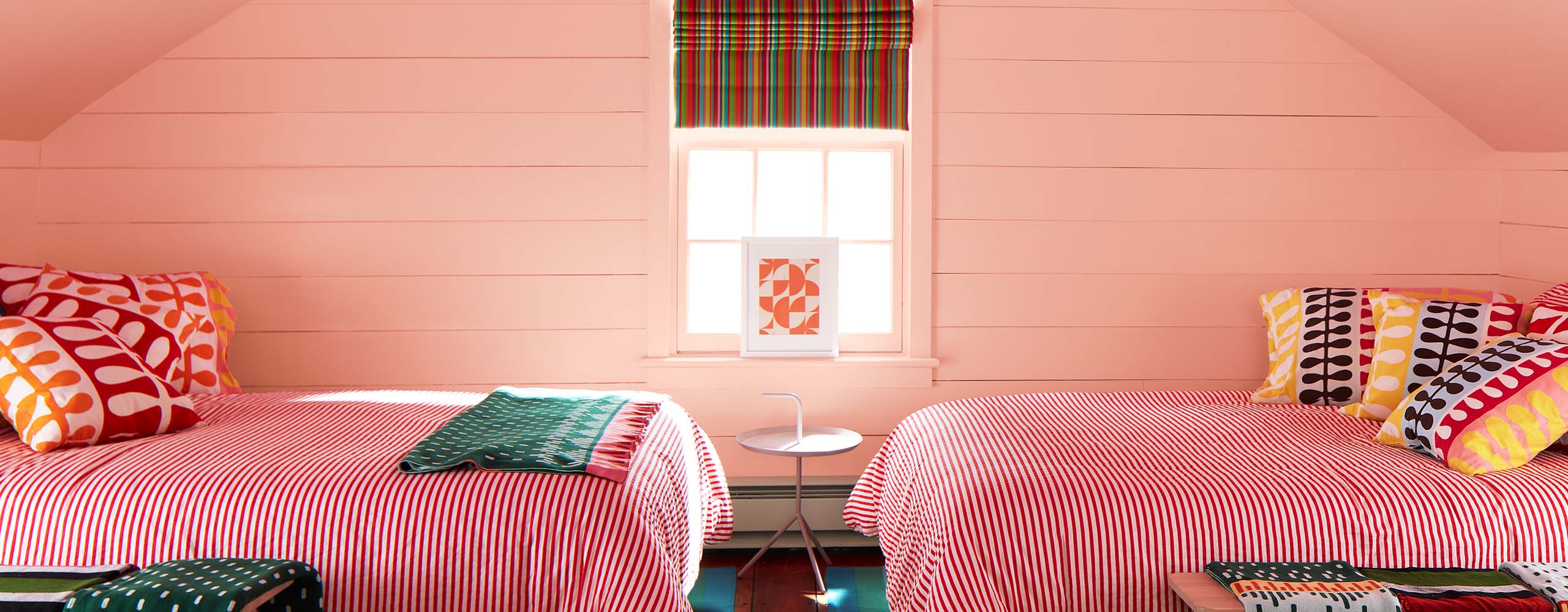 A bedroom painted a pink-orange pastel hue on the shiplap walls and vaulted ceiling features colorful bedding and decor, mixing stripes and patterns.