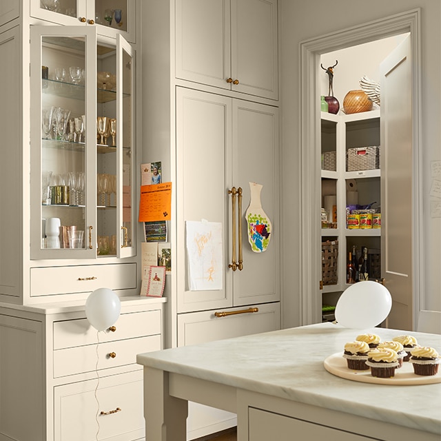 A bright, beige-painted kitchen with floor-to-ceiling and glass cabinets, an island with a marble countertop, an open pantry door, and white balloons on the floor and island.
