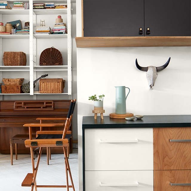 Bright, white-painted loft space with black-painted upper cabinets, a two-tone white and wood lower cabinet, a director’s chair, and tall white shelves in a back room over a piano.