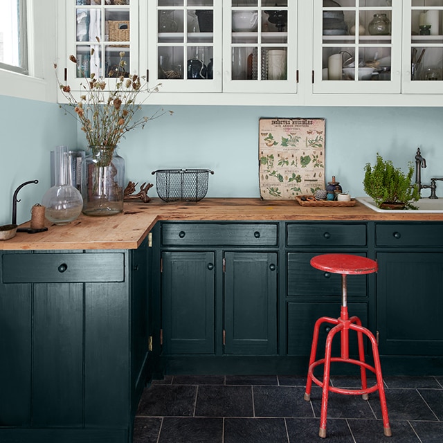 A modern farmhouse kitchen with light blue painted walls, white cabinets, dark green-painted lower cabinets and a red stool.