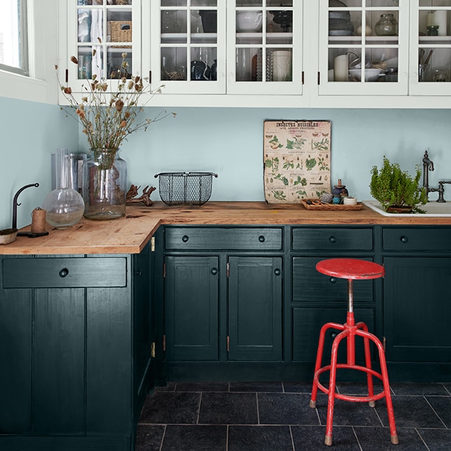 Cocina de casa rural moderna con paredes pintadas de azul claro, gabinetes blancos, gabinetes inferiores pintados de verde oscuro y un banco rojo.