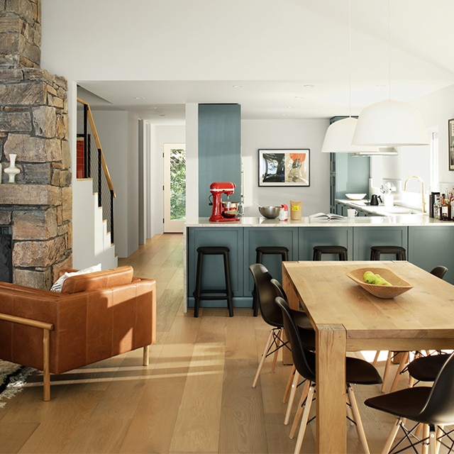 An open floor plan living space with white-painted walls and vaulted ceiling, dark gray kitchen cabinets, a wooden dining table and black chairs, and stone fireplace.