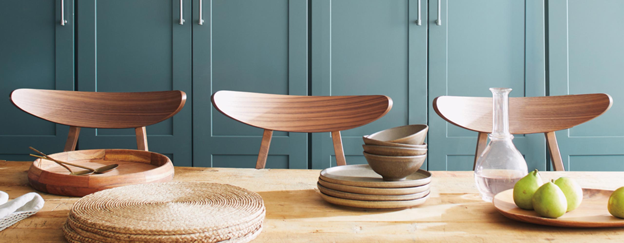 A wall of beautiful light teal-painted kitchen cabinets behind a wooden tabletop with wicker placemats, wooden tableware, and three wood chairs.