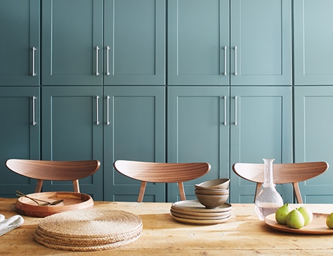 A wall of beautiful light teal-painted kitchen cabinets behind a wooden tabletop with wicker placemats, wooden tableware, and three wood chairs.
