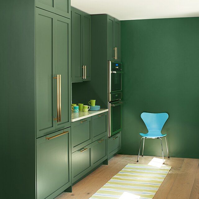 A sleek kitchen space with uniformly green painted built-in cabinets and wall, a white ceiling, modern turquoise chair, and a yellow-striped rug on a blonde wood floor.