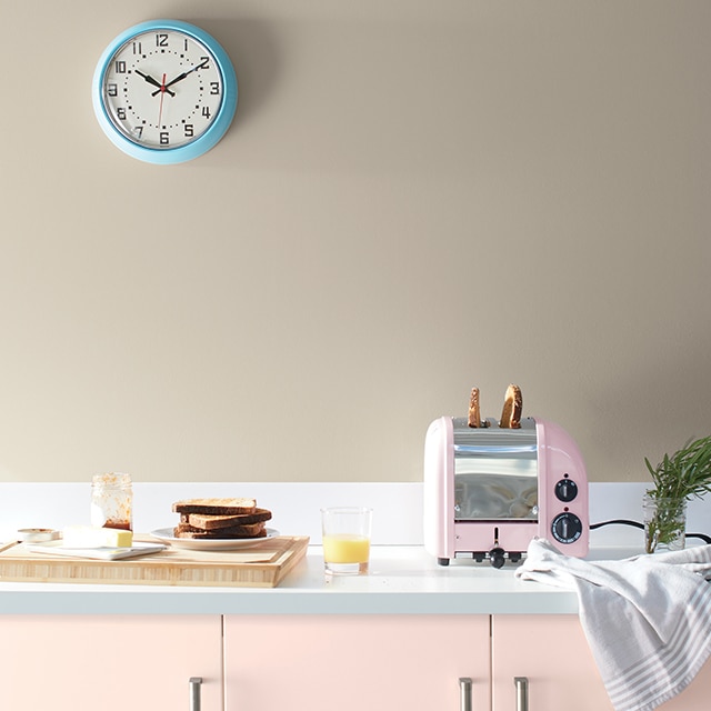 Una cocina con gabinetes pintados de rosa claro, una tostadora rosa, una tabla para cortar, platos y una pared pintada de bronceado con un reloj azul.