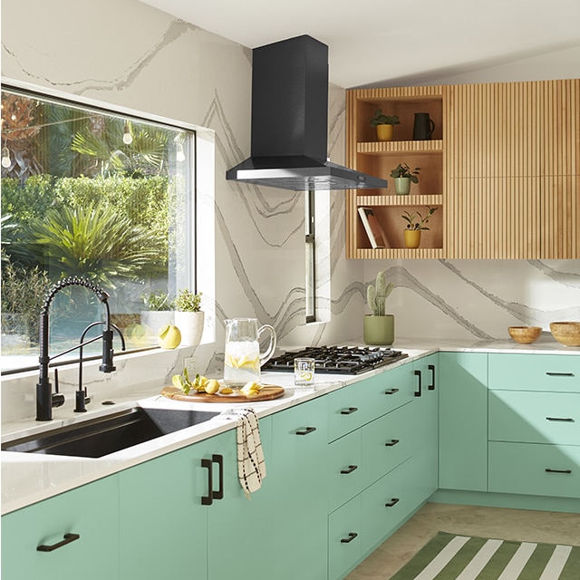 An airy white kitchen with lower cabinets painted in a light mint green, a large picture window over a black sink, white countertops, and upper cabinets with open shelving in light wood. 