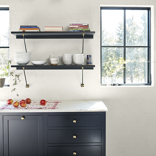 A pretty sunlit kitchen with dark navy-painted cabinets and floating shelves, a white wall, and natural wood beamed ceiling. 