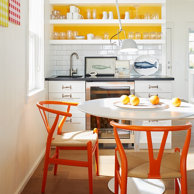 A white-painted kitchen with white cabinets and open shelving, a yellow accent wall, a white table and orange chairs.