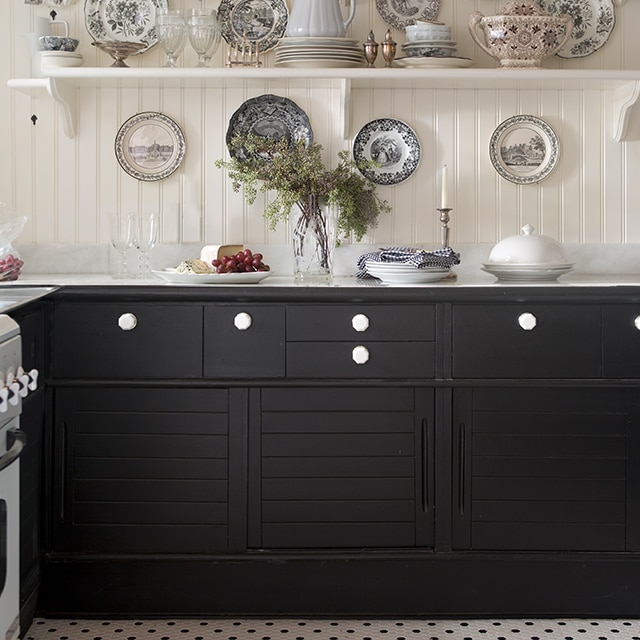 Una cocina al estilo campiña francesa pintada de blanco con estanterías abiertas y platos estampados arriba de gabinetes pintados de negro, y un piso de mosaicos blanco y negro.