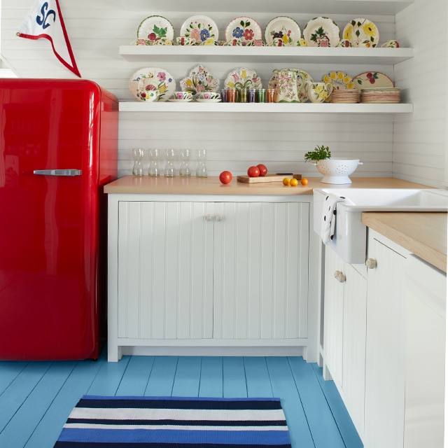 Una cocina pintada de blanco con suelo pintado de azul claro, refrigerador rojo y estanterías flotantes blancas.