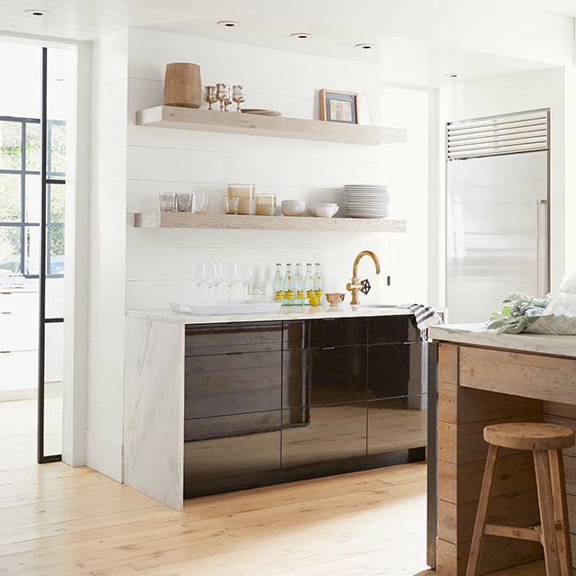 A pleasant, white-painted kitchen with black cabinets, marble countertops, open wood shelving holding tableware, and a wood-based island and stool.