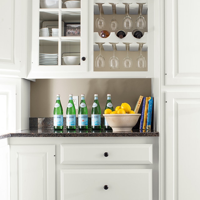 White-painted kitchen cabinets, a built-in wine rack and drawers, with a black countertop displaying water bottles and a bowl of lemons. 