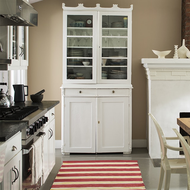 A greige-painted kitchen wall with a tall white hutch, white fireplace, white kitchen cabinets, and a red and white striped rug.