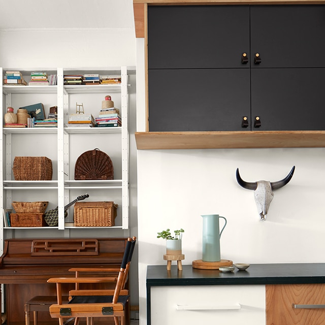 Una sección pequeña de una cocina abierta exhibe gabinetes y cajones pintados en negro y blanco con estantes decorativos y una silla de director alta más allá.