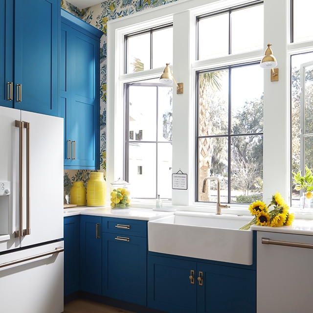 A sunny kitchen with blue-painted cabinets, white ceiling and trim, floral wallpaper, white appliances and a bunch of sunflowers.