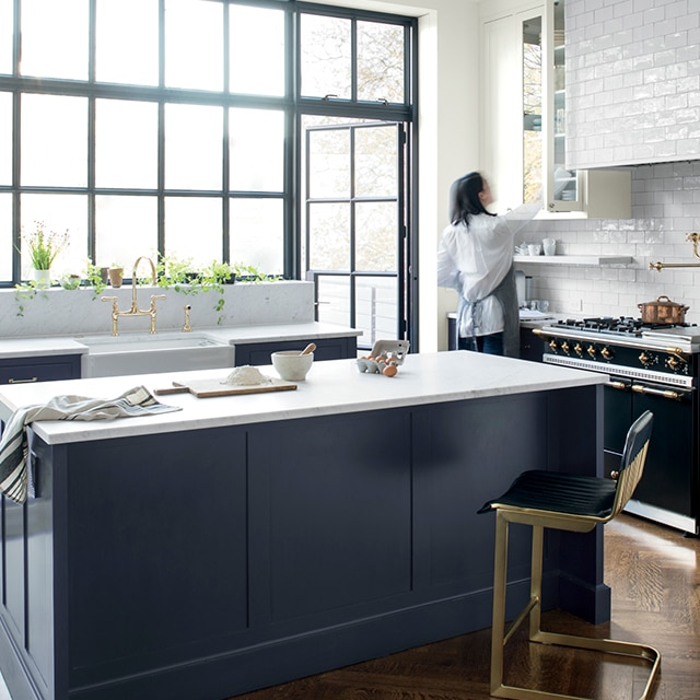 Cuisine lumineuse à aire ouverte avec grandes fenêtres et porte vitrée, îlot et armoires du bas bleu marine, comptoir blanc, armoire suspendue vitrée blanche et dosseret à carreaux blancs.