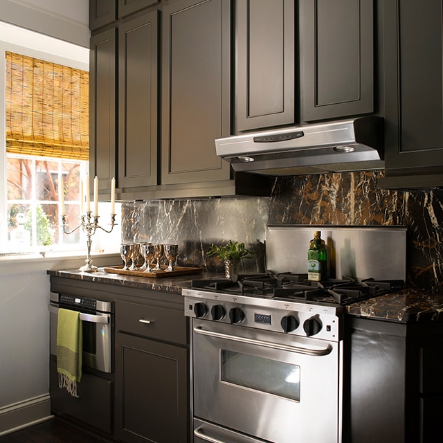 Rich, brown-painted kitchen cabinets, a brown marble backsplash and stainless-steel stove next to a white wall with a window and bamboo shade.