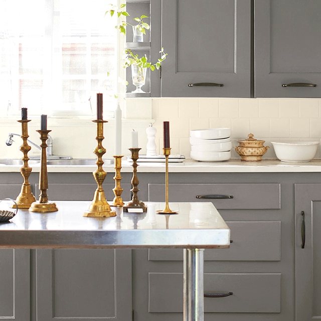 A traditional kitchen with dark gray-painted cabinetry, off-white tile backsplash, a white-trimmed window, and a stainless-steel worktable set with gold candlesticks.