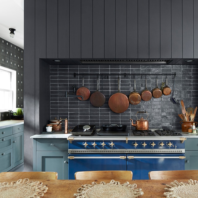 A warm, inviting kitchen with blue-painted cabinets, a black shiplap wall and black backsplash, a white ceiling, blue stove, and wood table and chairs.