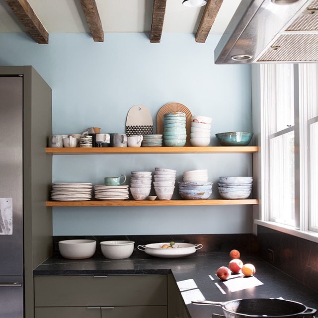 Cuisine rustique avec mur d’accent bleu pâle, armoires du bas vert mousse foncé, étagères suspendues en bois où s’entassent des bols et des assiettes, et comptoir noir.