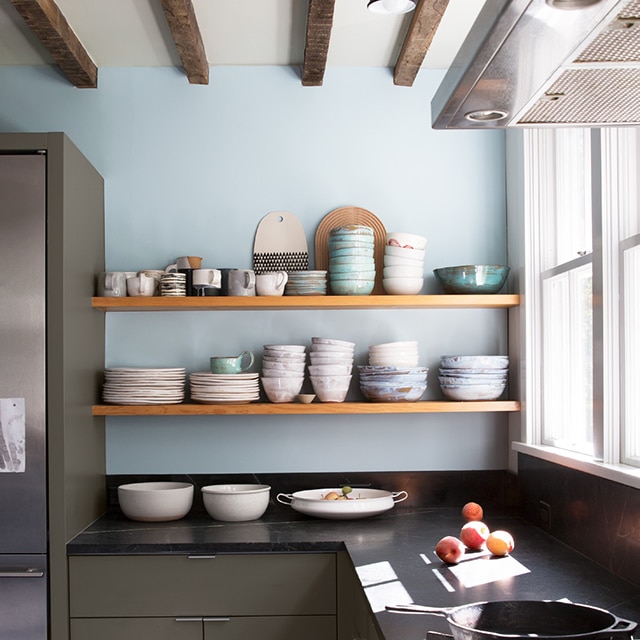 Una cocina rústica con una pared de acento pintada de azul claro, gabinetes inferiores pintados de verde musgo oscuro, estanterías de madera abiertas con boles y platos apilados, y una encimera negra.