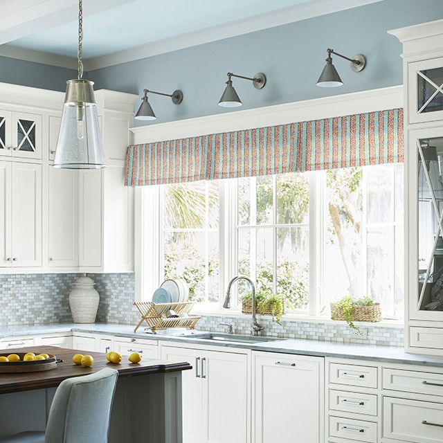 Charmante cuisine avec armoires blanches, murs du haut bleu clair, plafond bleu pâle à poutres blanches et cantonnière à rayures rouges le long des fenêtres.
