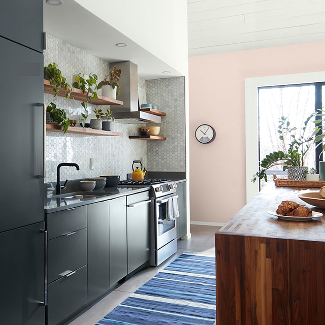 Una cocina pequeña acogedora con gabinetes pintados en gris carboncillo, protector contra salpicaduras de azulejos en gris perlado, una pared de acento en rosa y una alfombra a rayas azules.